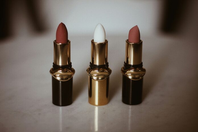 Three lipsticks with gold and black cases featuring lip-shaped accents, displayed on a smooth surface. Shades include two darker tones and a white.